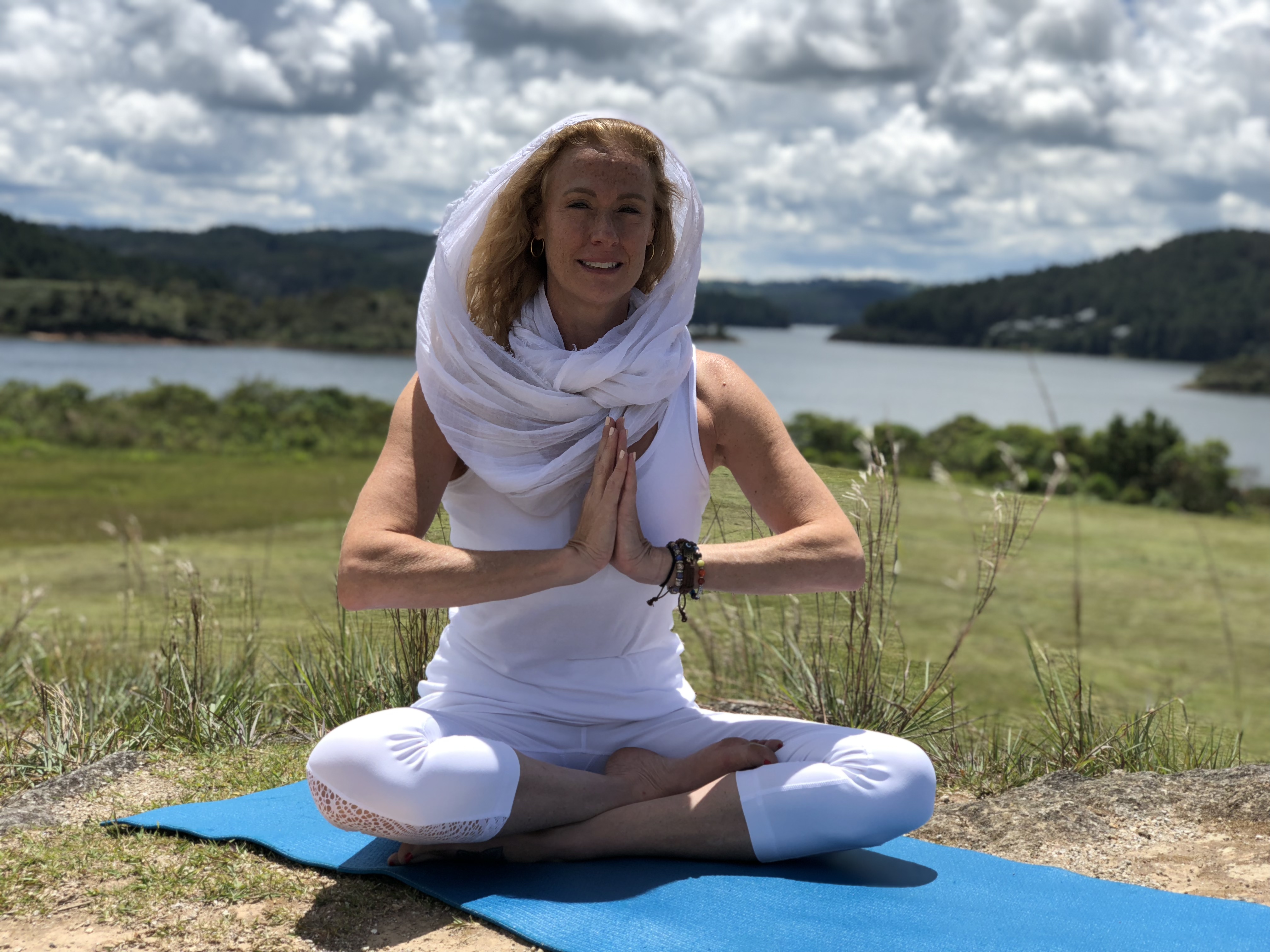 VictoriAnn in white, seated in prayer pose outdoors by a lake