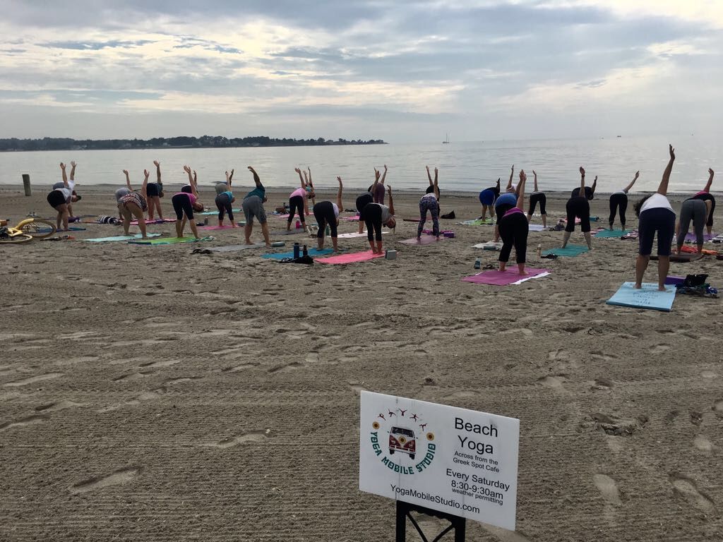 Una clase de Yoga Mobile Studio en una playa, todos con un brazo estirado sobre la cabeza en un estiramiento lateral junto al agua