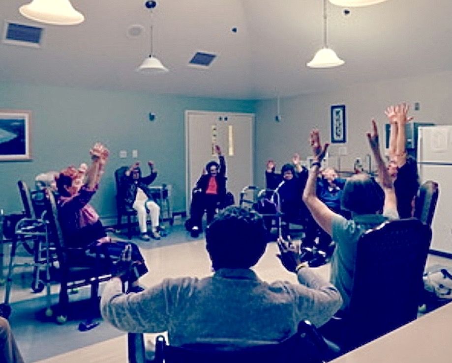 A seated group class in a bright common room, everyone reaching both arms overhead from their chairs