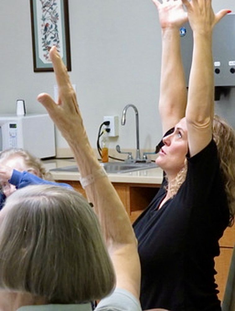 VictoriAnn with her arms stretched high, guiding a seated stretch while students in front of her do the same