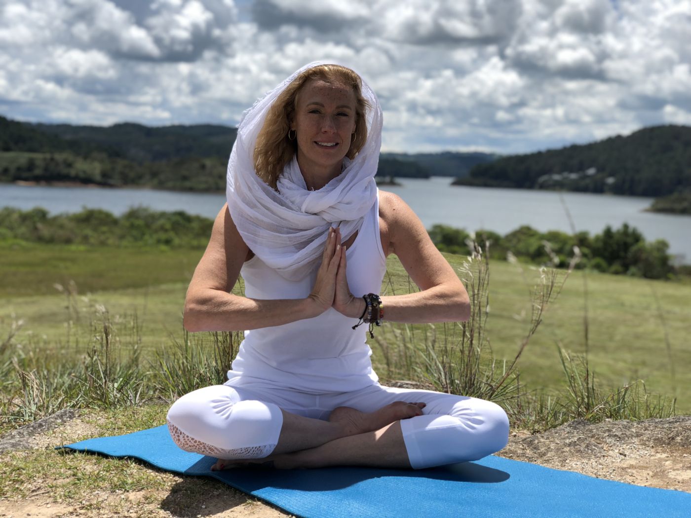 VictoriAnn smiling in a seated prayer pose on a blue mat, wrapped in a white scarf, with a lake and green hills behind her